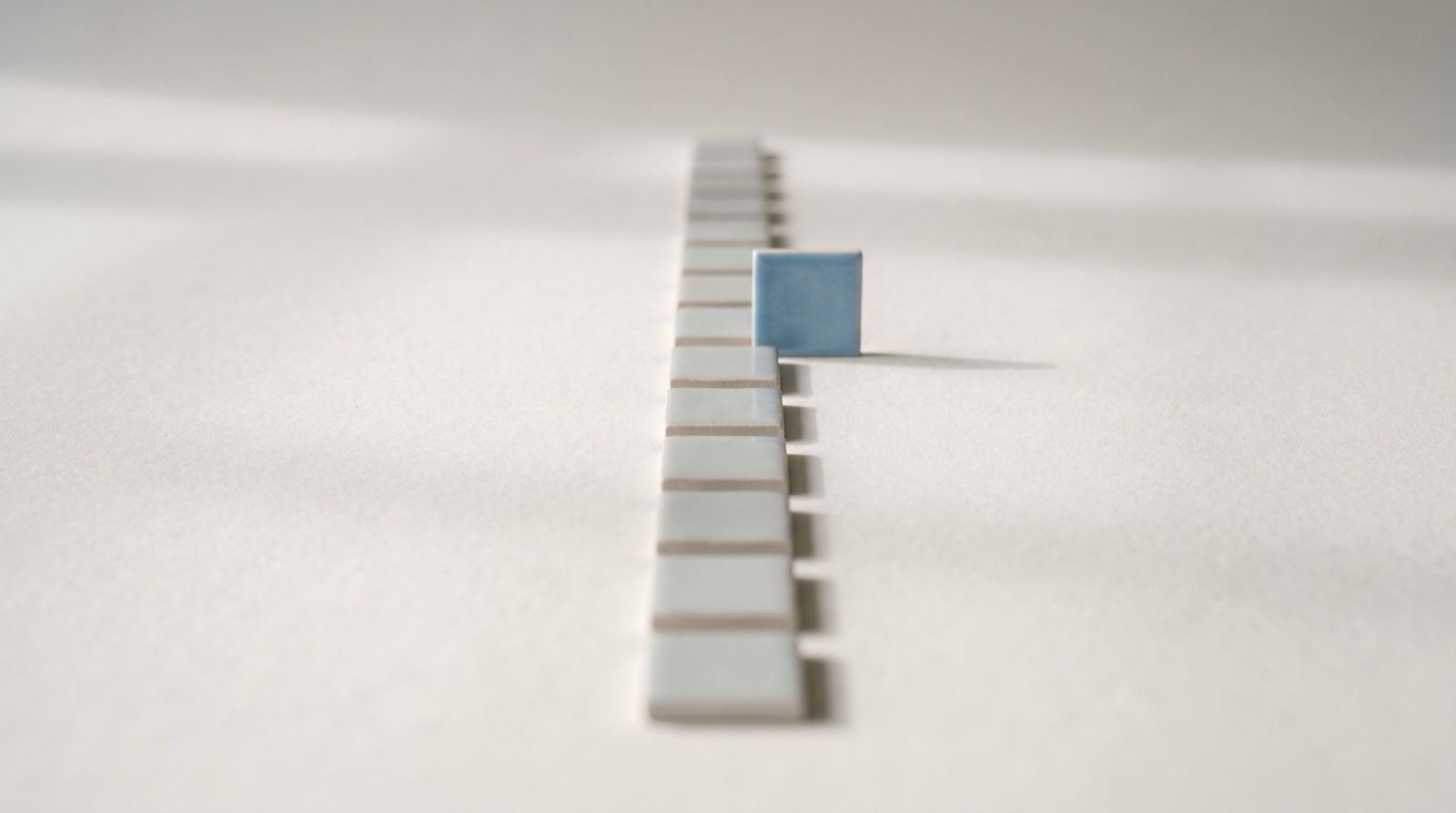 A line of small pale-grey tiles laid out like stepping stones across an off-white surface, with one sky-blue tile standing upright partway along the path where it will be met.