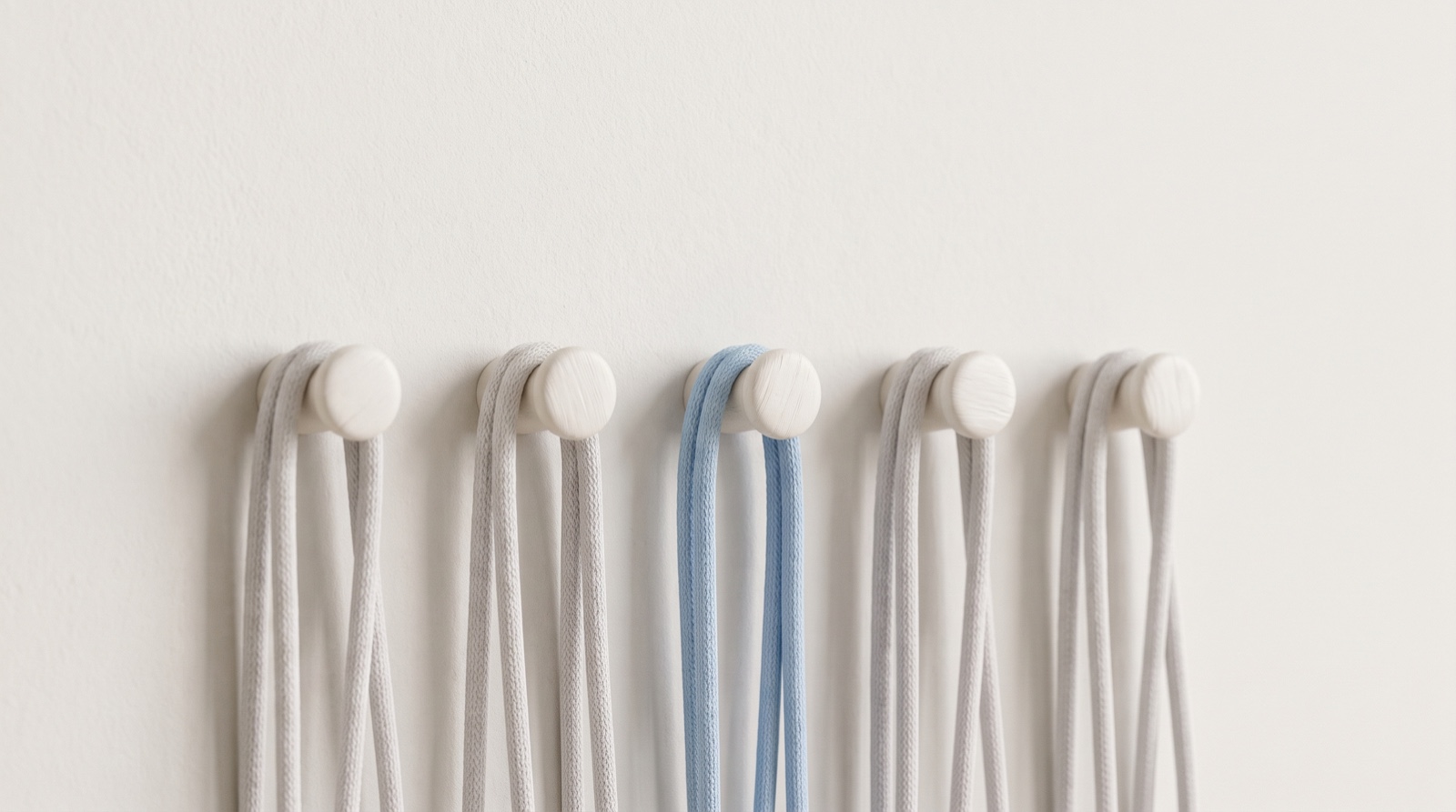 A row of five small round pegs on a calm off-white wall, each holding hanging pale-grey cords, with one set of cords in sky-blue, like small fixes holding the day in place.