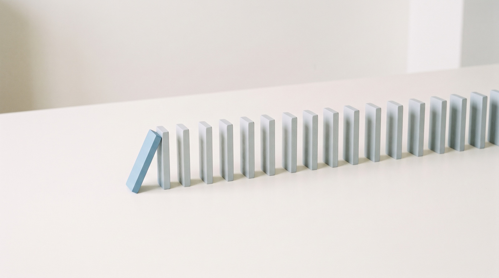 A long row of upright pale-grey tiles on an off-white surface, with the first tile in sky blue tipped over and resting against the next one.