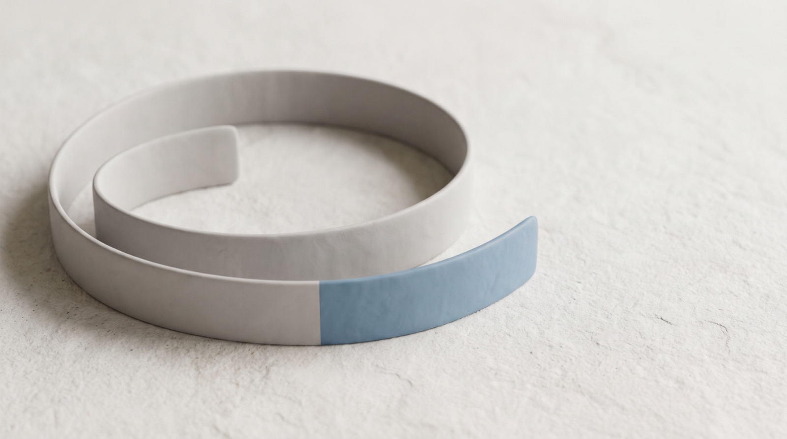 A smooth pale-grey band coiled in a loose spiral on a calm off-white surface, one short segment of it in sky-blue, like a stretch of time made visible enough to measure.