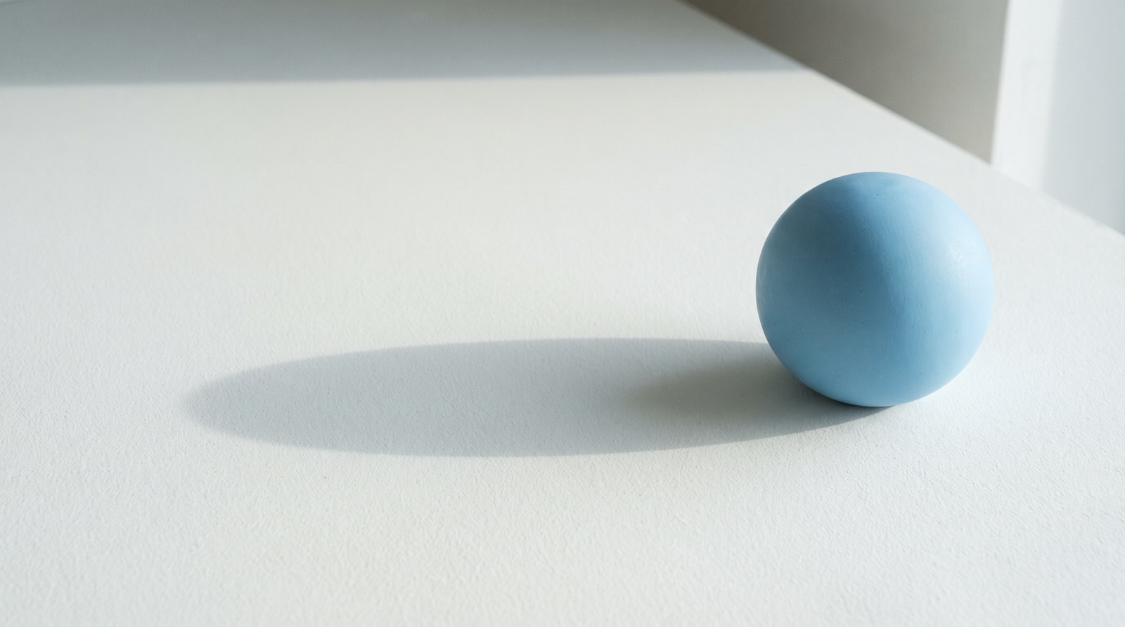 One small sky-blue sphere near the right edge of a wide off-white surface, its long soft shadow stretching back across the empty space before it.
