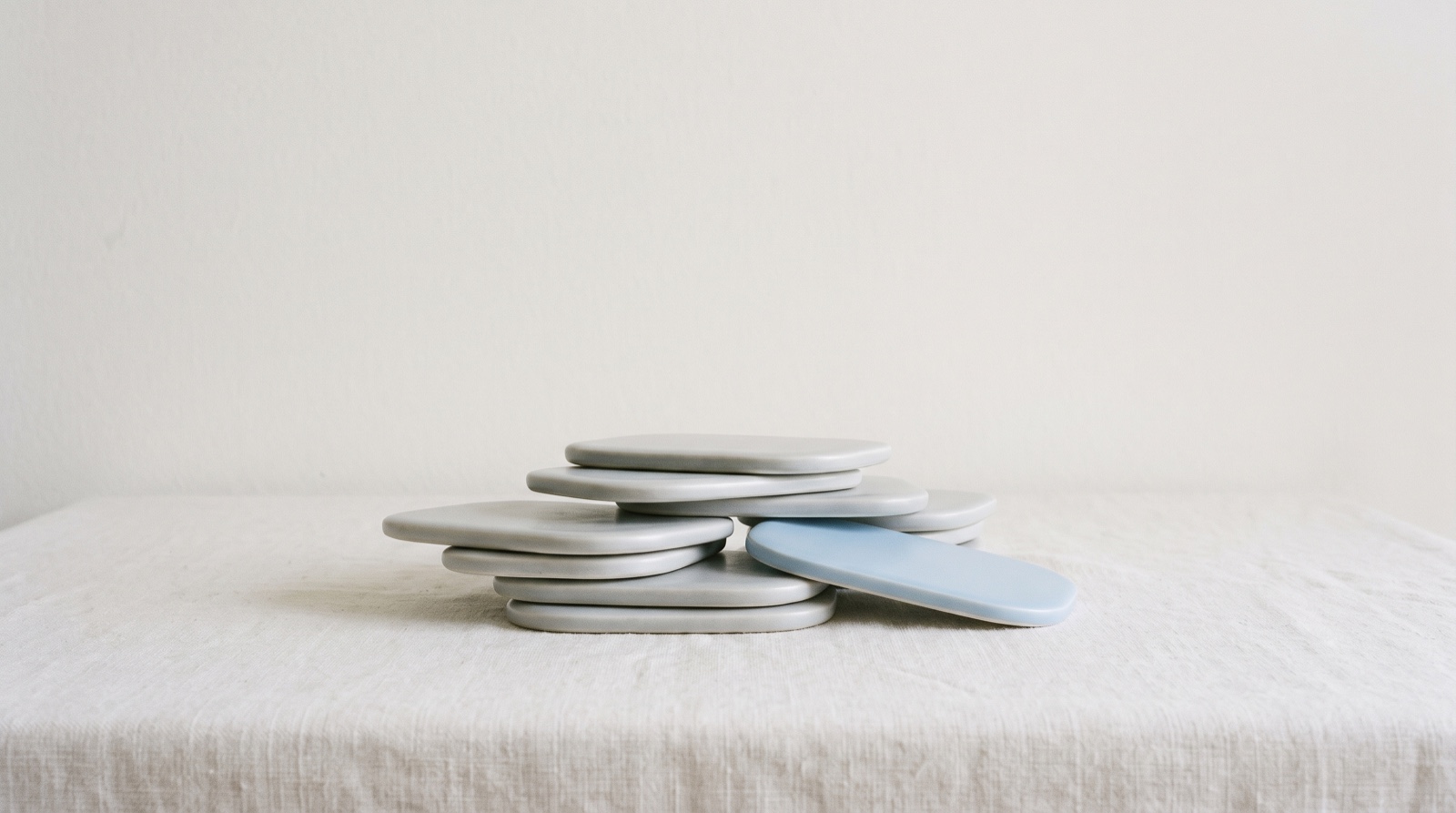 A small, loosely stacked pile of soft pale-grey tiles on a calm off-white surface, with a single sky-blue tile resting among them.