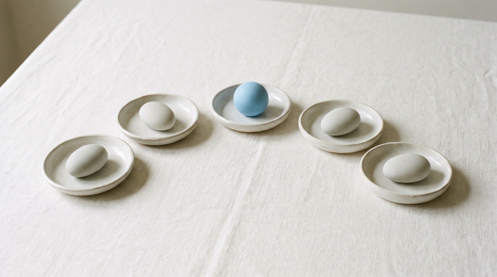 Five shallow pale ceramic dishes set in a gentle arc on a calm off-white cloth, each holding one smooth stone, with the center dish holding a single sky-blue sphere.