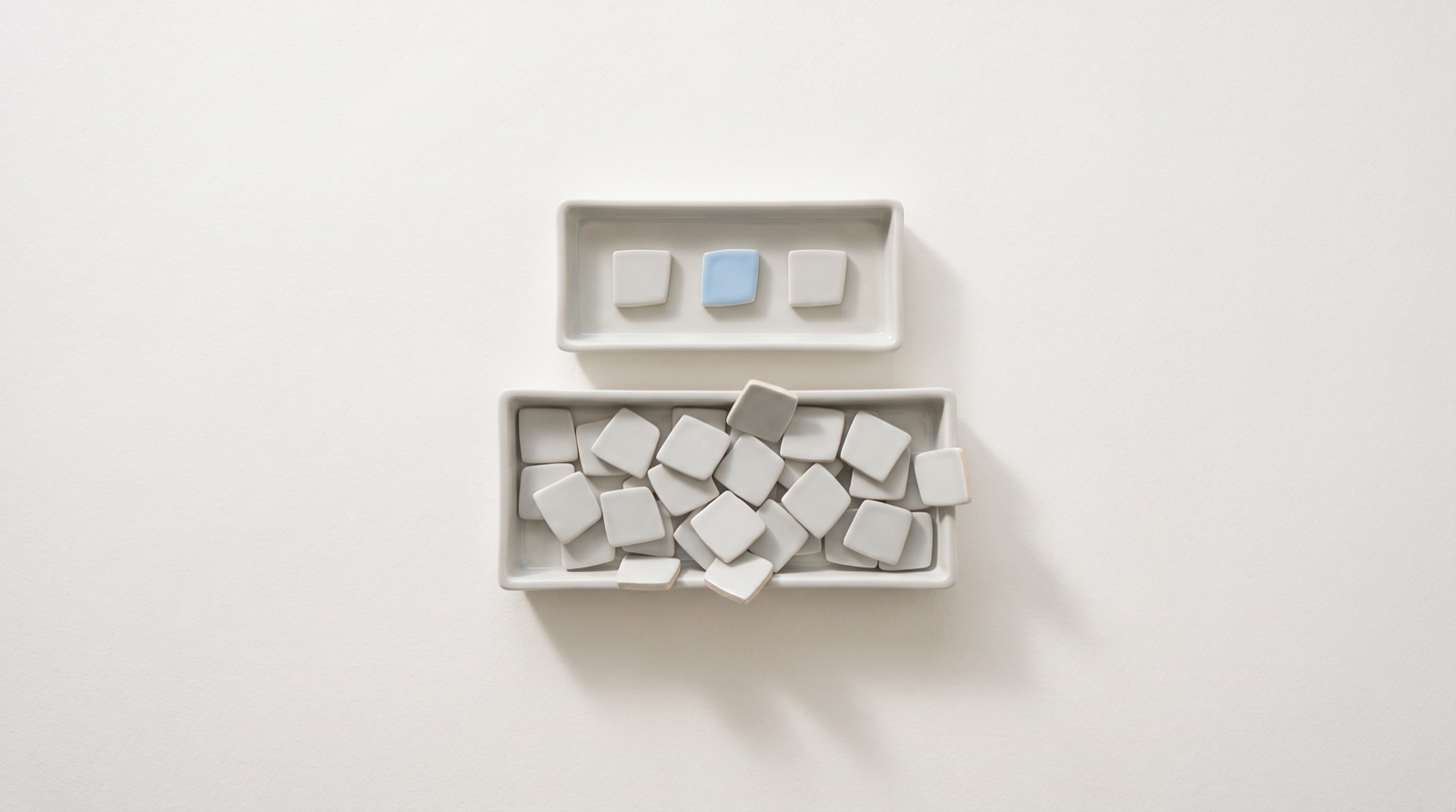 Two pale ceramic trays on a matte off-white surface, the lower one heaped with small grey tiles and the upper one holding just three in a row, one of them sky-blue.
