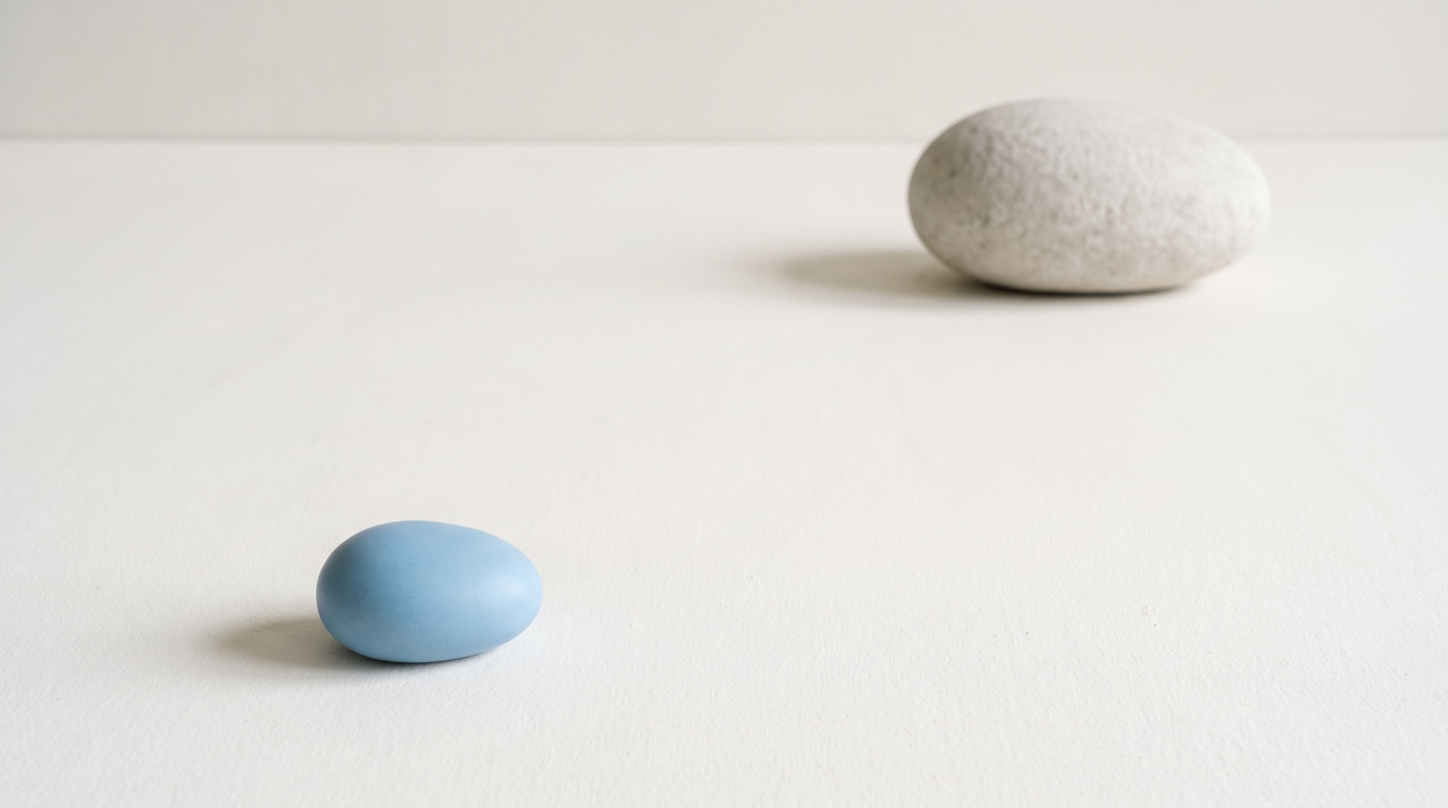 A small smooth sky-blue pebble resting close to the viewer on a matte off-white surface, with a much larger rough pale-grey stone sitting far back across the empty space.