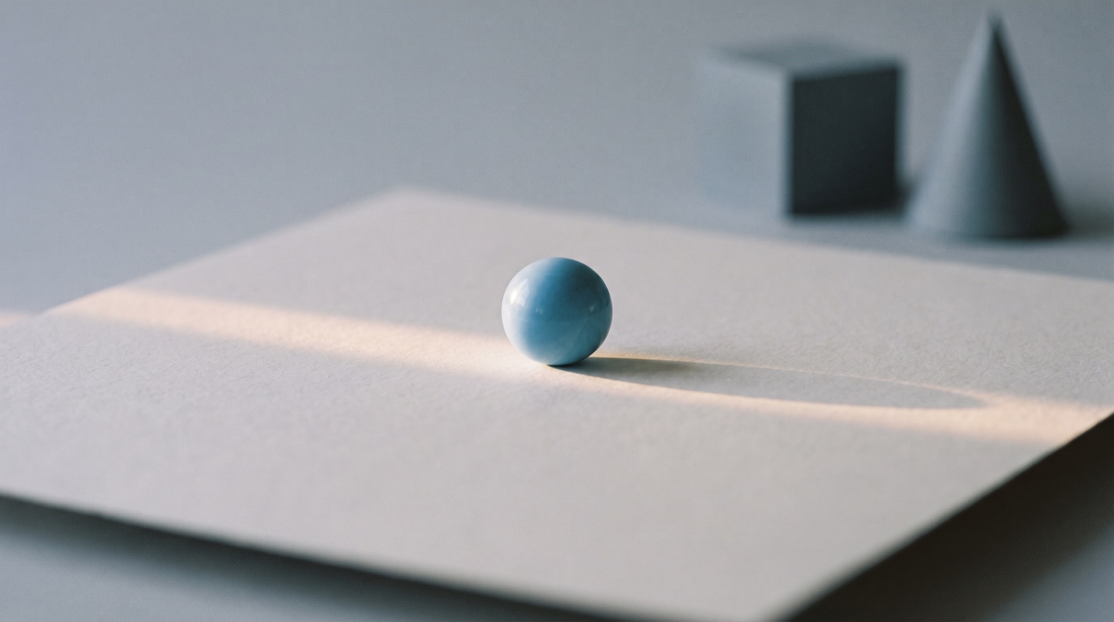 A single sky-blue sphere resting in a narrow band of light on a matte off-white surface, with a few pale-grey shapes sitting out of focus in the dimmer background.