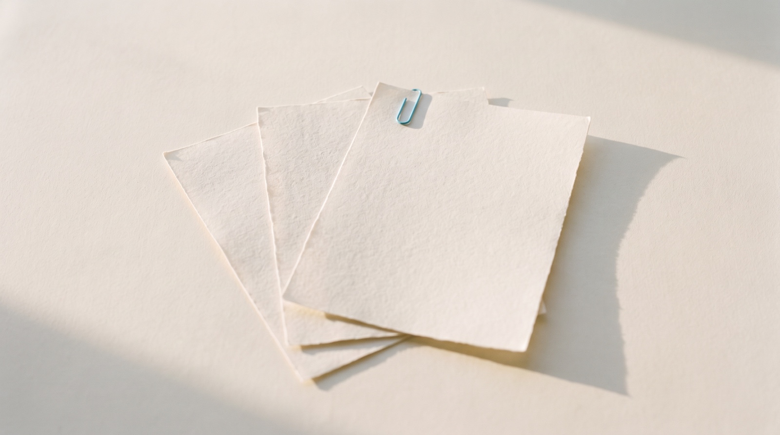 Three blank sheets of pale paper fanned in a soft stack on an off-white surface in low morning light, one small sky-blue clip on the top sheet, like a quiet page waiting before the day starts.
