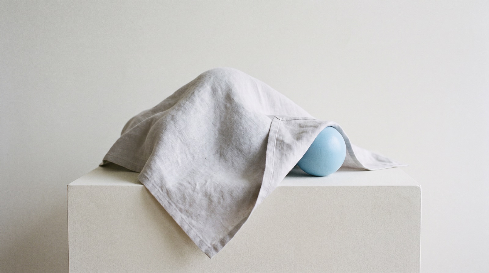 A soft pale-grey cloth draped over a small form on a calm off-white surface, with a single sky-blue sphere emerging into view at the edge of the cloth.