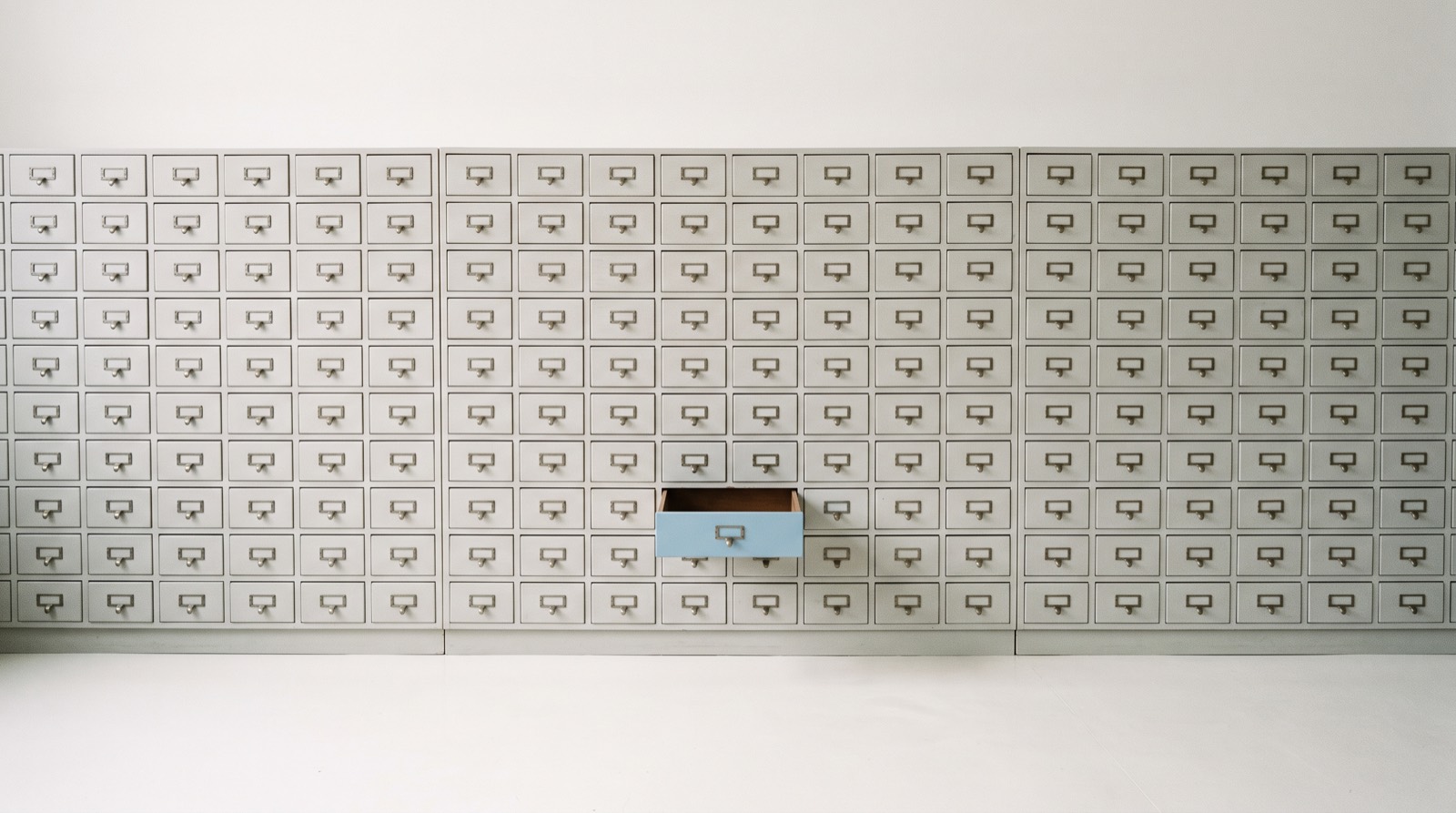 A wide wall of small closed pale-grey drawers on a matte off-white surface, with one sky-blue drawer pulled open, like a single idea retrieved from a large store.