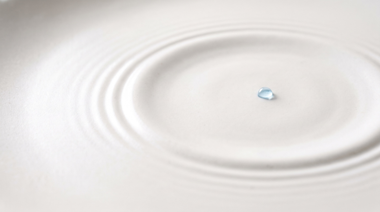 A single slow ripple spreading outward from one small sky-blue droplet across a wide, still off-white surface, with the rest of the frame left empty.