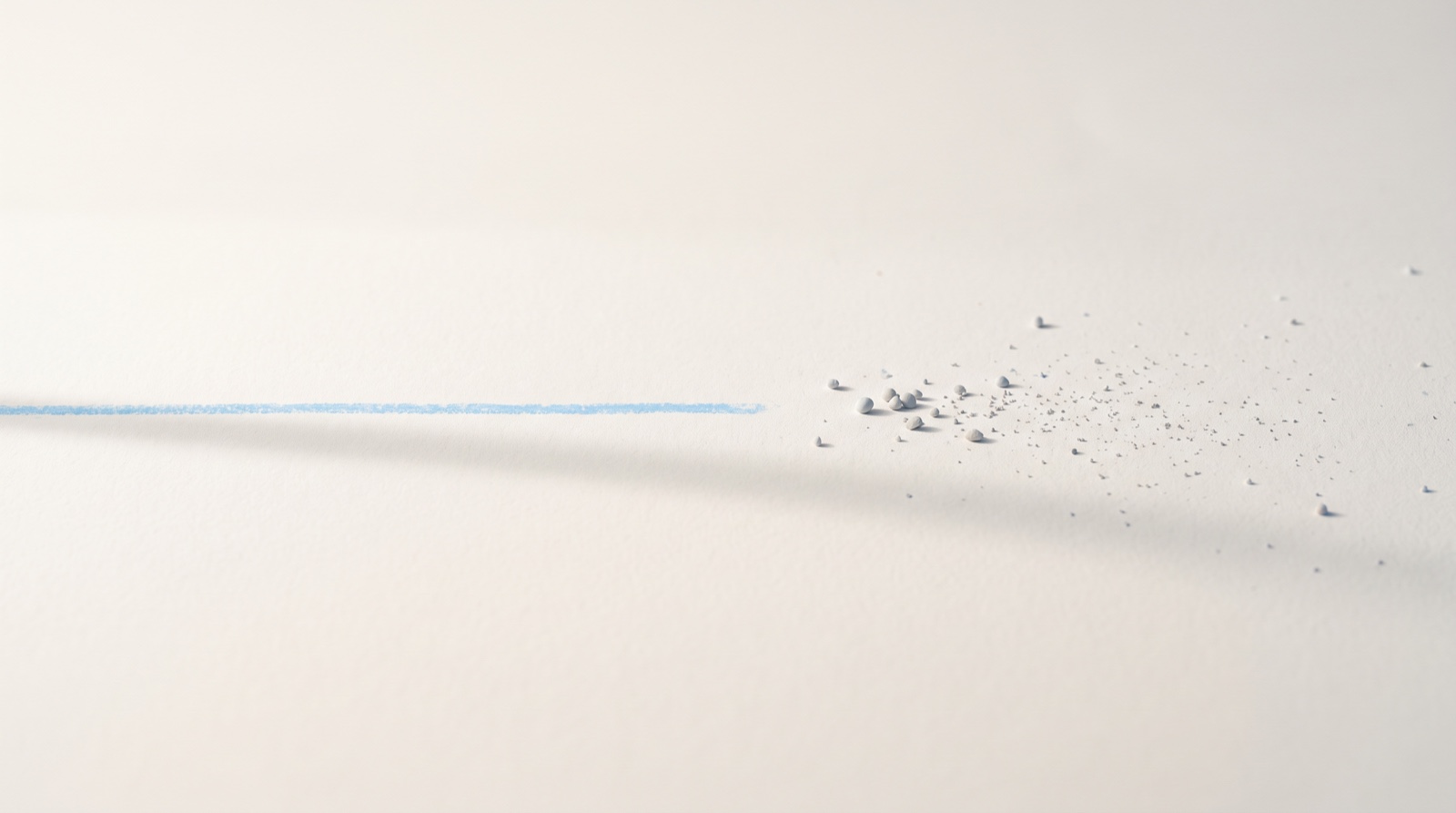 A slim sky-blue line drawn partway across a matte off-white surface, the area behind it smooth and empty, with a fine scatter of small pale-grey grains left beyond where the line ends.